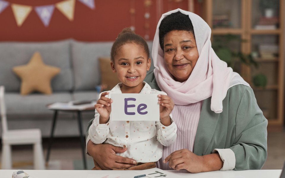 grootmoeder met hoofddoek zit trots naast kleindochter die letters toont in een klasje